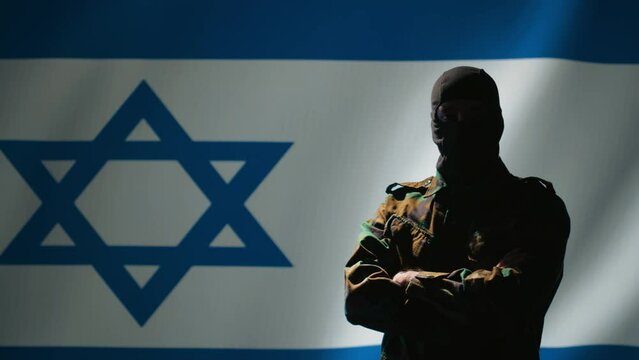 Mossad operator wearing mask to remain anonymous standing proud in front of Israel flag, feeling pride in serving country. Israeli secret agent in command room crossing arms, camera B
