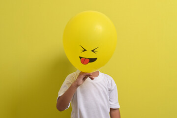 Tongue emoji. Cute Asian boy hiding his face behind balloon emoticon sticking out tongue isolated on yellow background. Teasing emoji with closed eyes