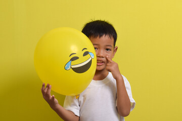 Laughing emoji. Cute Asian boy holding laughing emoticon balloon isolated on yellow background