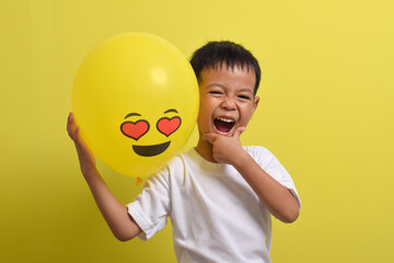 Emoticon in love. Cute Asian boy holding romantic love face emoticon balloon isolated on yellow background