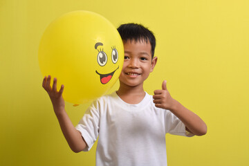 Smile emoji. Cute Asian boy holding happy emoticon balloon isolated on yellow background