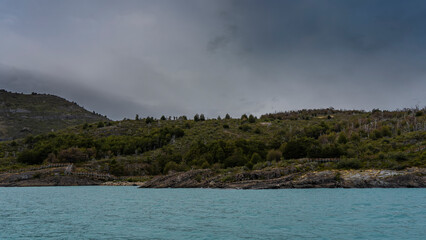 Fototapeta premium Turquoise glacial lake. There is a forest growing on the coastal hills. The layered structure of the rocks is visible. Clouds in the sky. Lago Argentino. El Calafate. Argentina. Los Glaciares 