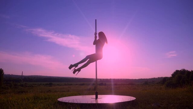 Silhouetted woman in a swimsuit gracefully pole dancing in a sunlit field. Elegant pole dancer performing at twilight in an open field. Beautiful sunset backdrop as woman dances on a pole in meadow. W