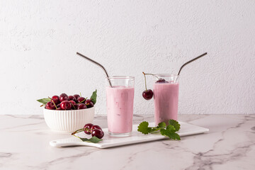 Two tall glasses of cherry smoothie on a white ceramic tray and a bowl of ripe, juicy cherries. marble countertop. A white brick wall. dessert. Snack.