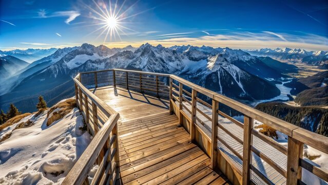  There is a wooden walkway with a railing on top of a mountain. The walkway is surrounded by snow. There are mountains in the distance. The sun is shining.