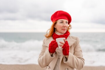 Obraz premium Woman beach in red beret scarf and mitts holding a cup of tea in his hands. Depicting beach relaxation and cozy attire. Walks by the sea