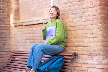 Happy smiling young man holding a notebook and pointing thumb up looking at camera at the university campus