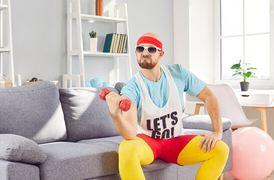 Young man wearing sunglasses, headband and T shirt with Let's go text having sports workout at home, sitting on sofa in living room, doing exercise with fitness dumbbell with funny facial expression