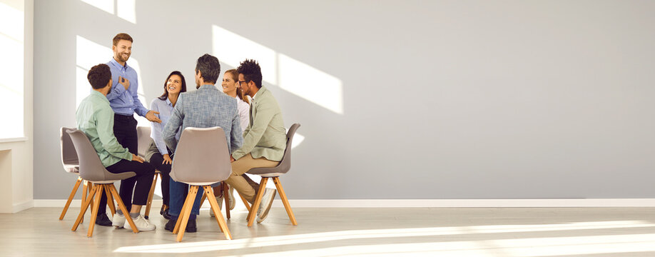 Business coach and team mentor meeting with group of people. Smiling man talking to happy young diverse men and women sitting in circle in modern office with gray copy space wall. Banner background