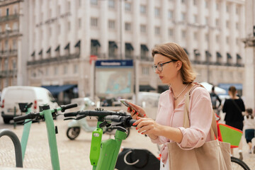 Middle aged woman renting an electric scooter or bicycle using smartphone, making contactless payment through mobile app. City travel and solo tourism concept. Copy space. Part of the series