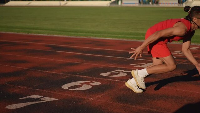A male athlete in red sportswear, sunglasses and dreadlocks assumes the crouch start position and starts running forward.