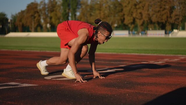 Slow motion. An athlete in scarlet sportswear, sunglasses and dreadlocks takes position crouch start on the running track of the city stadium and after the start begins to run screaming