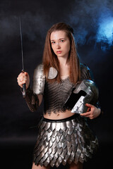 Portrait of a medieval female warrior in armor with a sword and helmet in her hands, standing in smoke with blue backlight isolated on a dark background.
