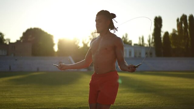 Slow motion. Shooting against the sun. At sunset, an athlete with dreadlocks in red gym shorts jumps with a jump rope on a running track against the green grass of the stadium field. Training jumping
