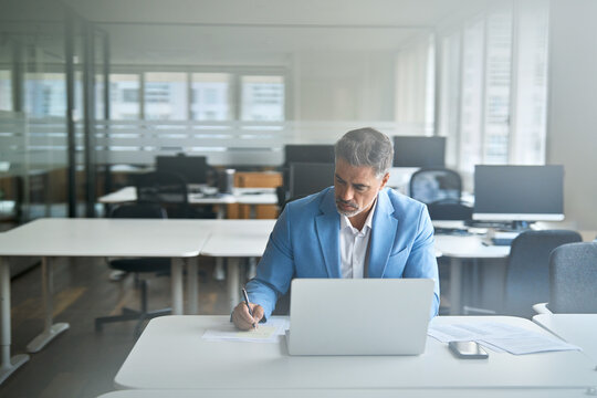 Busy older middle aged professional business man executive manager wearing suit working on financial analysis using laptop computer in office writing notes. View through glass. Candid photo.