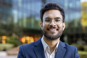 Confident young man with glasses and beard wearing suit smiling outdoors in urban setting. Business professional expressing optimism and readiness. Background features blurred office buildings