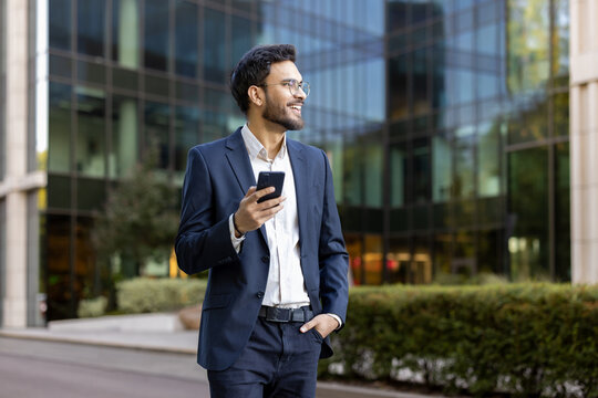 Confident young businessman in suit holding smartphone and smiling while standing outdoors near modern office building. Professional attitude, business success, and positive outlook convey success