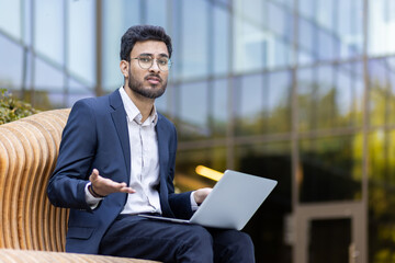 Confused businessman sitting on bench outdoors using laptop. Professional dressed in suit shrugging shoulders, perplexed expression. Modern glass building background. Concept of business problem