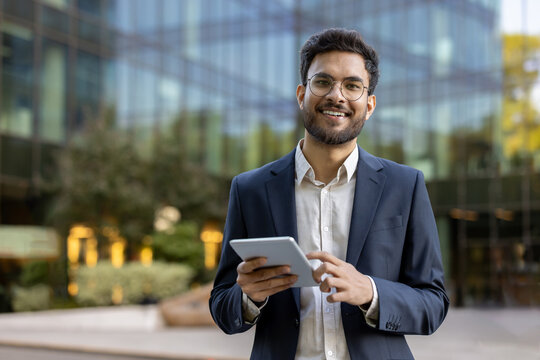 Young businessman in suit using tablet outside modern glass office building. Confident professional smiling working on digital device. Concept of business, technology, success, and entrepreneurship.