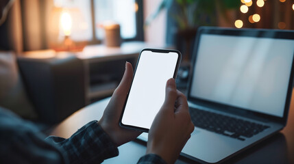 Person holding smartphone with blank screen next to laptop in cozy home setting