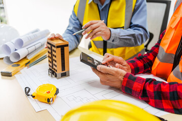 Two men, including a male Asian construction contractor engineer, discuss structural plans and engineering concrete columns and beams of a new house at a desk with a building tower model.