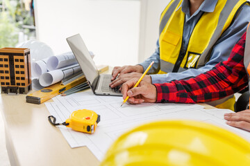 Two men, including a male Asian construction contractor engineer, discuss structural plans and engineering concrete columns and beams of a new house at a desk with a building tower model.