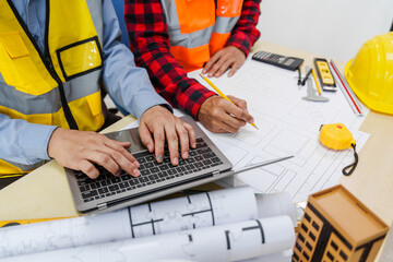 Two men, including a male Asian construction contractor engineer, discuss structural plans and engineering concrete columns and beams of a new house at a desk with a building tower model.