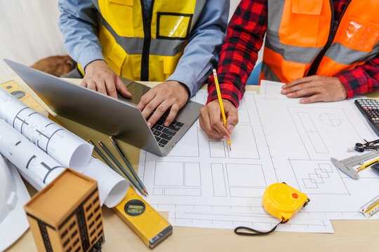 Two men, including a male Asian construction contractor engineer, discuss structural plans and engineering concrete columns and beams of a new house at a desk with a building tower model.