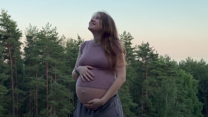Young happy pregnant woman walking in the pine forest at summer day and stroking her big belly 