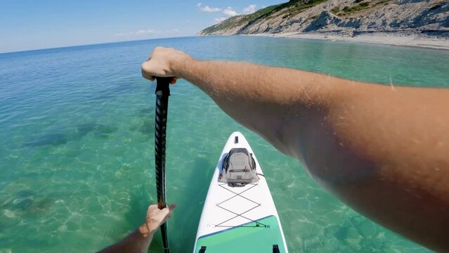 First person view of stand paddle boarding on quiet turquoise water. Men rowing on sup board on summer sea.