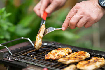 Man grilling flipping meat on barbecue grill grid in his backyard. close up of man making barbecue in his backyard, a man uses tongs to flip a grilled meat on a barbecue grill, green grass in the back