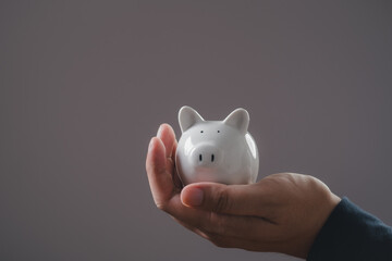 Close-up of a hand holding a white piggy bank against a gray background, signifying savings,...