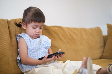 Fototapeta premium Young child sitting on a couch, concentrating on a smartphone with a serious expression, in a cozy indoor setting.