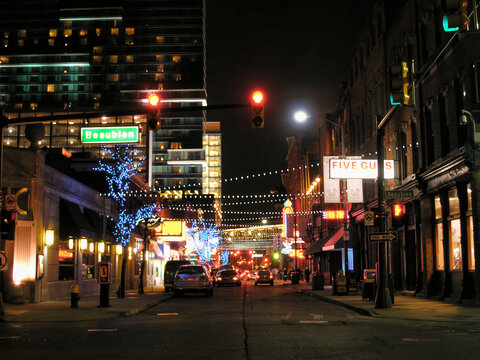 A vibrant nighttime scene in Detroit, Michigan. Street is illuminated by colorful lights and decorations. Storefronts and businesses visible.