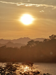 Stunning sunset landscape featuring a golden sun illuminating a serene mountain lake. Misty peaks and tree silhouettes create a tranquil atmosphere, with warm light reflecting off calm waters.
