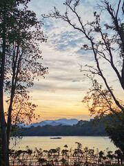 Serene lakeside sunset: silhouetted trees frame golden sky over mountains. Lone boat on calm waters embodies tranquility, natural beauty, and harmony between man and nature.