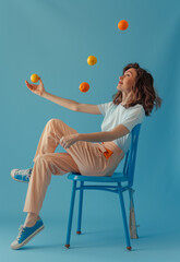 Fototapeta premium A woman sitting on a blue chair, juggling balls and a peach in the air.Minimal creative fun concept.