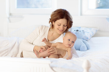 Mother and child on a white bed. Mom and baby boy in diaper playing in sunny bedroom. Parent and little kid relaxing at home. Family having fun together. Bedding and textile for infant nursery.