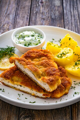 Crispy breaded seared chicken cutlet with boiled potatoes and cucumbers in cream served on white plate on wooden table