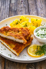 Crispy breaded seared chicken cutlet with boiled potatoes and cucumbers in cream served on white plate on wooden table