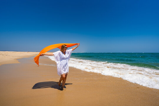 Summer vacation. Beautiful young woman in white tunic and sunglasses holding orange scarf fluttering in the wind walking on the sandy beach by the sea with big waves on a beautiful sunny day