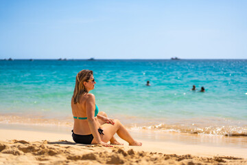 Beach vacation. Happy young woman in bikini sitting on sandy beach by the sea on beautiful sunny day. Positive emotion