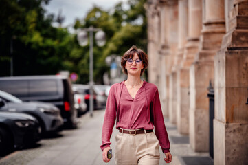 Woman in casual attire smiling while walking on a city sidewalk lined with parked cars and buildings.