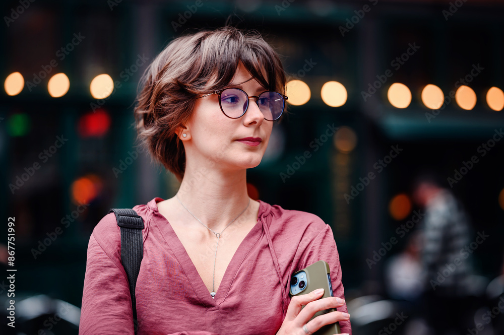 Poster Close-up of a confident woman with glasses holding her phone, standing in front of an urban background with lights.