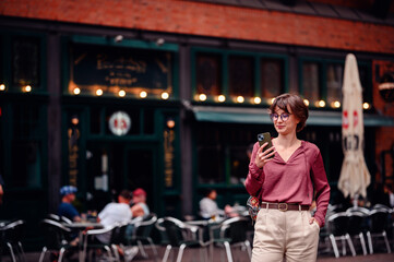 Obraz premium Woman in casual attire checking her phone while standing outside a busy cafe with people seated at tables.