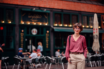 Fototapeta premium Woman in casual attire smiling and holding her phone while standing outside a cafe with people sitting at tables.
