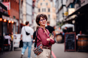 Smiling woman in casual attire and glasses with a colorful backpack, standing on a busy city street lined with cafes and shops.