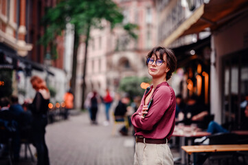 Obraz premium Woman in casual attire and glasses, smiling confidently with her phone and backpack, standing in a bustling city street lined with cafes and people.