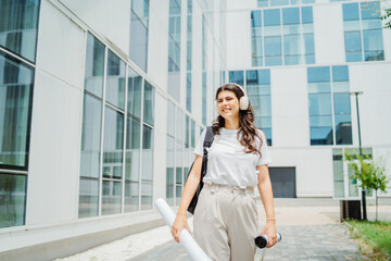 Fototapeta premium One young caucasian woman student going to university while listening to music on wireless headphones and holding papers and coffee 
