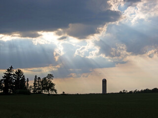 Trees and a lone silo are silhouetted by the sun beaming through the clouds. 